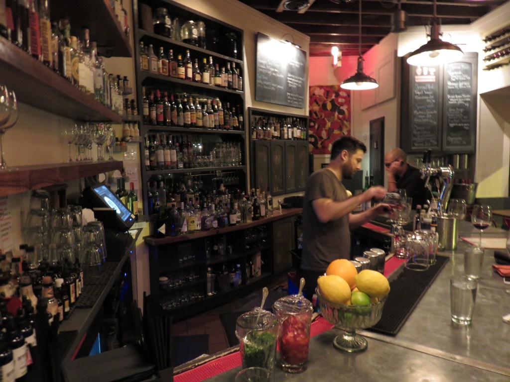 Photograph of the interior of a bar. Glassware and gruit line the far edge of the bar. A bartender is pouring a drink a few seats down. In the background is large dark shelf full is liquor bottles.