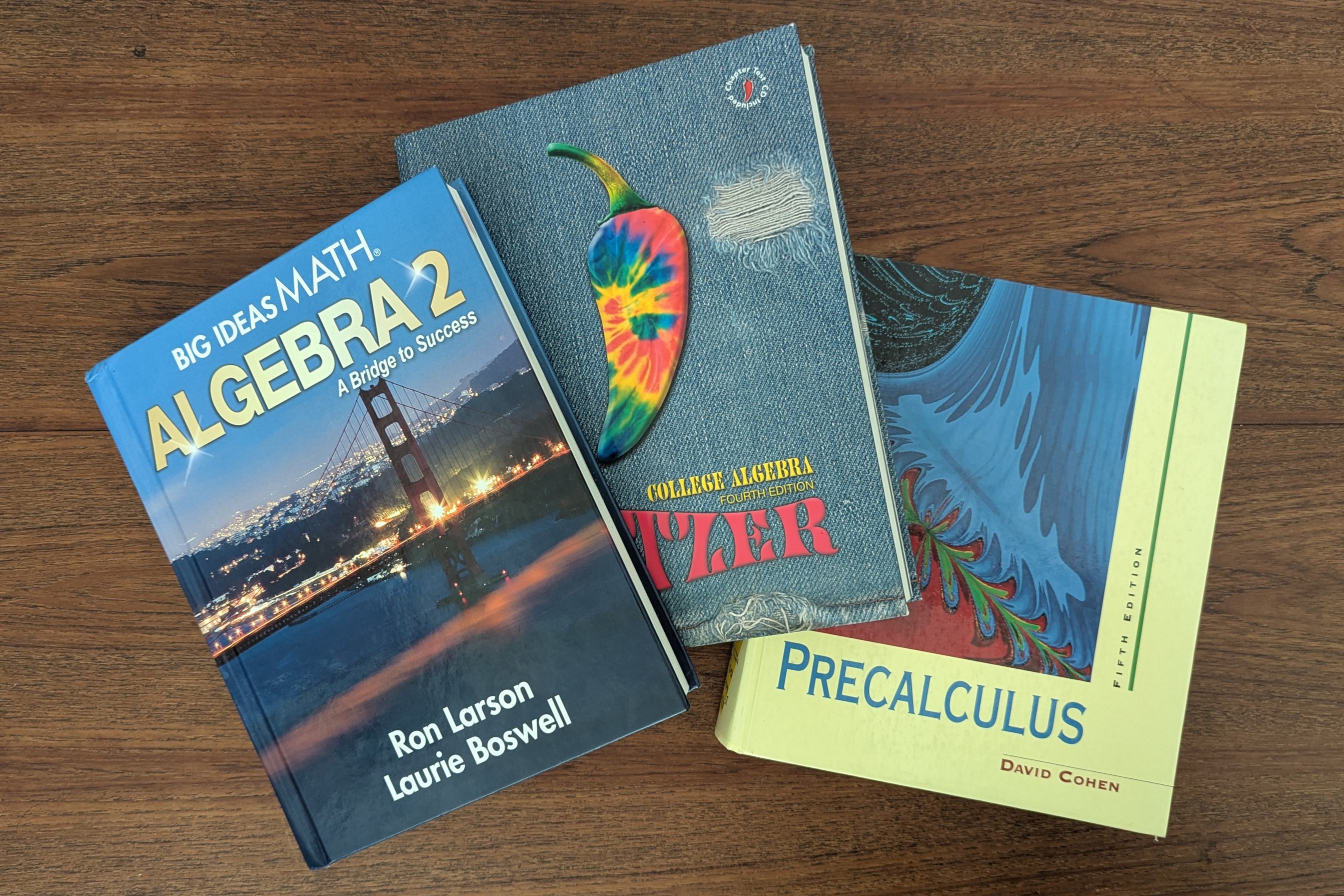 A photograph of three textbooks fanned out on a wooden table. They are Algebra 2 by Ron Larson and Laurie Boswell, College Algebra by Robert Blitzer and Precalculus by David Cohen.
